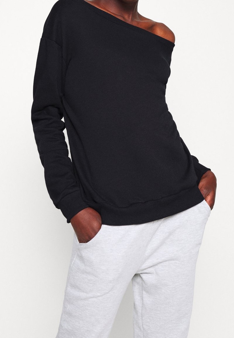 Sweat-shirt noir à épaules dénudées en tissu doux, avec une coupe décontractée et des poignets côtelés, assorti à un pantalon de survêtement gris clair.