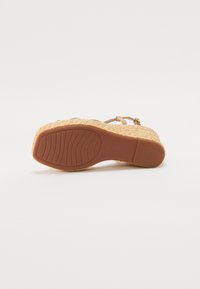Hælrem sandal med en flettet stråplattform og tan gummisåle. Har en tynn stropp over foten og justerbar ankelstropp.