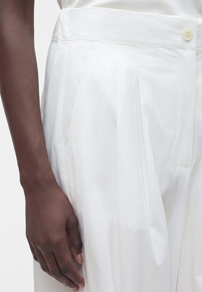 Main à la peau foncée reposant à côté d'un pantalon blanc taille haute avec des plis et un bouton visible à la taille.