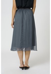 Femme portant une jupe midi semi-transparente gris charbon avec une taille élastique et des mules noires à talons, debout devant un fond uni.