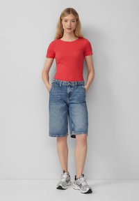 Czerwony prążkowany top z krótkim rękawem w połączeniu z jasnoniebieskimi dżinsowymi bermudami o prostym kroju i postrzępionym wykończeniu, noszony z srebrnymi butami sportowymi.