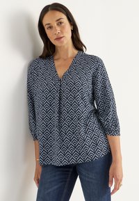 Blaue gemusterte Bluse mit V-Ausschnitt und Dreiviertelärmeln, die ein geometrisches Blättermuster in Hellblau und Marineblau zeigt. Weicher Stofftextur.