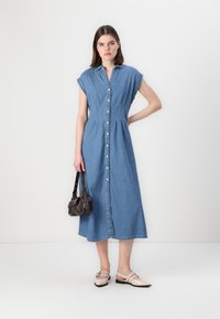 Abito midi in denim con maniche corte, chiusura a bottoni e dettagli plissettati. Indossato con scarpe beige e una piccola borsa scura.