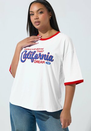 Frau mit langen schwarzen Haaren, die ein weißes T-Shirt mit rotem Rand und blaue Jeans trägt. Auf dem Shirt steht der Text „Woke up with that California Dream 1987.“