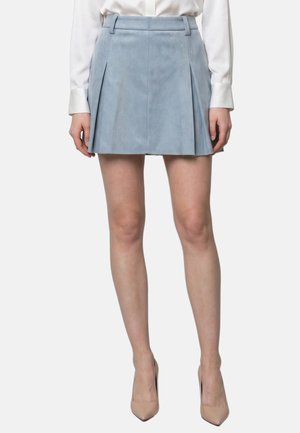 Donna con gonna plissettata azzurra, camicetta bianca a maniche lunghe e scarpe beige con tacchi alti, in piedi su sfondo uniforme.