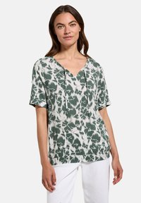 Top corto a maniche corte verde e bianco con scollatura a V, caratterizzato da un motivo a foglia e stringhe regolabili sul collo. Tessuto leggero.