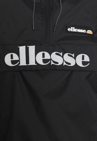Sort vindjakke med en kvart lynlås. Har et bredt sølvfarvet "ellesse" logo på brystet samt et ekstra mærkat.