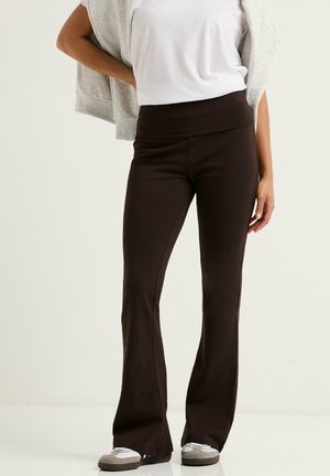 Kvinde iført sorte vidde bukser, hvid t-shirt, grå sweatshirt lagt over skuldrene og hvide sneakers, står mod en ensfarvet baggrund.