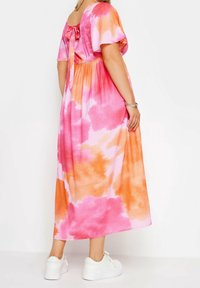 Maxi šaty v růžovo-oranžovém tie-dye vzoru, s krátkými rukávy, čtveratým výstřihem na zádech s mašlí a volným střihem, doplněné bílými teniskami.