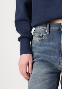 Marinblå sweatshirt med ribbade ärmslut, parat med ljusblå denimjeans som har en subtil blekning och en logotypetikett på frontfickan.