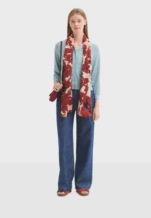 Jeune femme portant un cardigan bleu clair, un jean à jambes larges, une écharpe rouge à motifs floraux, des sandales rouges, et tenant un sac rouge à bandoulière en chaîne.