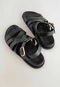 Des sandales en cuir noir présentent plusieurs larges brides et une seule bride ajustable avec une boucle en or rose. Texture lisse et design simple.