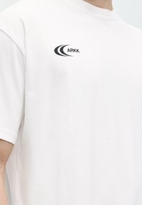 Λευκό μπλουζάκι που φοράει ένα άτομο, με μικρό μαύρο λογότυπο "ARKK" με καμπύλες γραμμές στην άνω αριστερή περιοχή του στήθους.