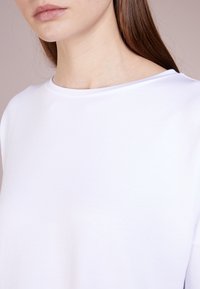 Detailní záběr na osobu v jednoduchém bílém tričku s kulatým výstřihem, s rovnými hnědými vlasy viditelnými přes jedno rameno.