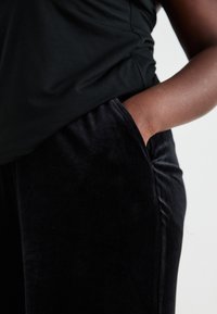 Pantalones de terciopelo negro con una textura suave, que cuentan con bolsillos laterales y un corte relajado, combinados con una blusa negra ajustada.