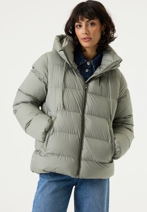 Femme aux cheveux foncés jusqu’aux épaules portant une doudoune gris clair sur une chemise en jean, les mains dans les poches, face à l’avant.