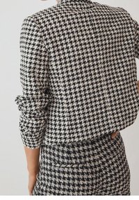 Femme portant une veste à motif pied-de-poule noir et blanc et un pantalon assorti, avec les manches légèrement froncées.