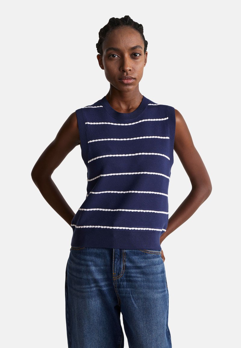 Granatowa bezrękawnikowa bluzka dzianinowa w poziome białe paski, okrągły dekolt, ściągacz na dole i luźny krój, w połączeniu z niebieskimi dżinsami.