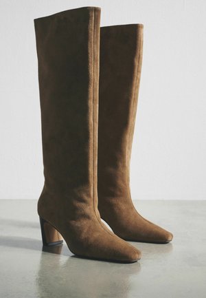 Hnedé semišové čižmy presahujúce kolená s špicatou špičkou a masívnym, uhlovým podpätkom. Hladká textúra a detailné švy.