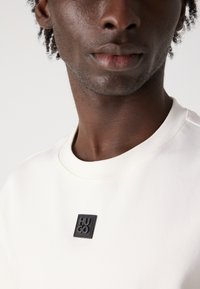 Helle Creme Sweatshirt mit rundem Ausschnitt, versehen mit einem schwarzen quadratischen Patch mit eingeprägtem "HUGO". Glatte Textur und minimalistisches Design.