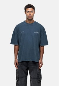 T-shirt en coton bleu surdimensionné avec un col rond et des manches courtes. Présente des bords effilochés et des accents de texte minimalistes à l'avant.