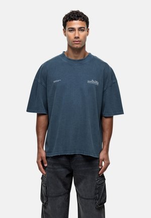 T-shirt blu di cotone oversize con scollo a giro e maniche corte. Presenta bordi sfilacciati e accenti testuali minimalisti sulla parte anteriore.