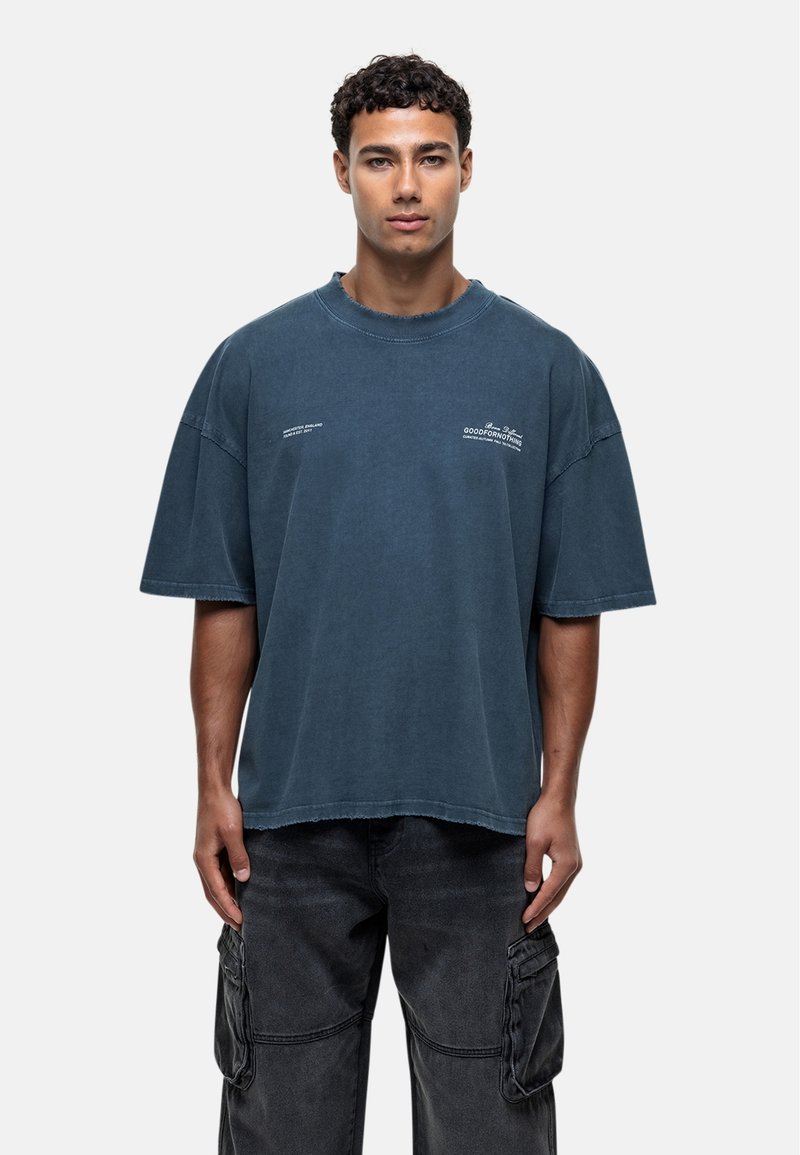 T-shirt en coton bleu surdimensionné avec un col rond et des manches courtes. Présente des bords effilochés et des accents de texte minimalistes à l'avant.