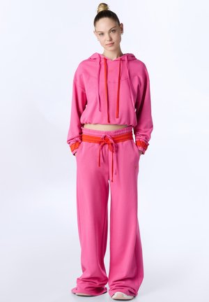 Donna in piedi con le mani nelle tasche, indossa una felpa corta rosa con cappuccio e pantaloni ampi abbinati con fascia in vita rossa e coulisse.