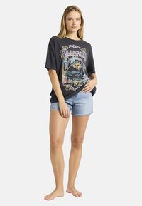 Μαύρο γραφικό μπλουζάκι με πολύχρωμο σχέδιο που περιλαμβάνει τη λέξη "BILLABONG 1973", σε συνδυασμό με ανοιχτό γαλάζιο τζιν σορτς, παρουσιασμένο σε ένα όρθιο άτομο.