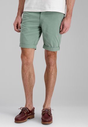 Man wearing rolled light green shorts, white shirt, and brown leather boat shoes standing on gray background.