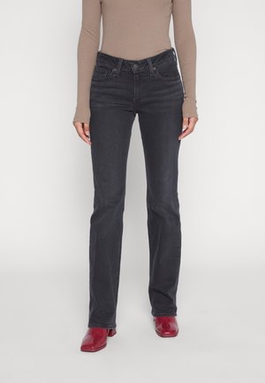 Personne portant un jean noir taille haute, un haut beige à manches longues et des chaussures à talons rouges, debout sur fond blanc.