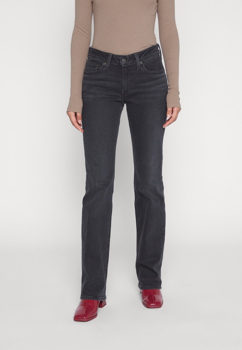 Personne portant un jean noir taille haute, un haut beige à manches longues et des chaussures à talons rouges, debout sur fond blanc.