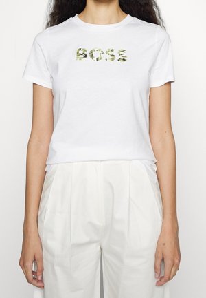 Nainen valkoisessa t-paidassa, jossa on "BOSS" vihreillä lehtikuvioisilla kirjaimilla, ja korkeavyötäröiset valkoiset laskostetut housut.