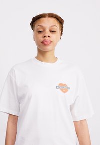 T-shirt en coton blanc avec un logo orange et bleu sur la poitrine gauche. Manches courtes et coupe décontractée. Texture lisse.
