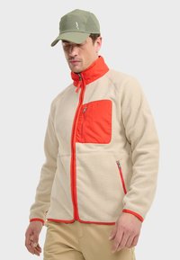Veste en polaire crème avec un col rouge et une poche sur la poitrine. Comprend des zips, des coutures contrastées et une coupe décontractée. Accentuée par une casquette en vert olive.