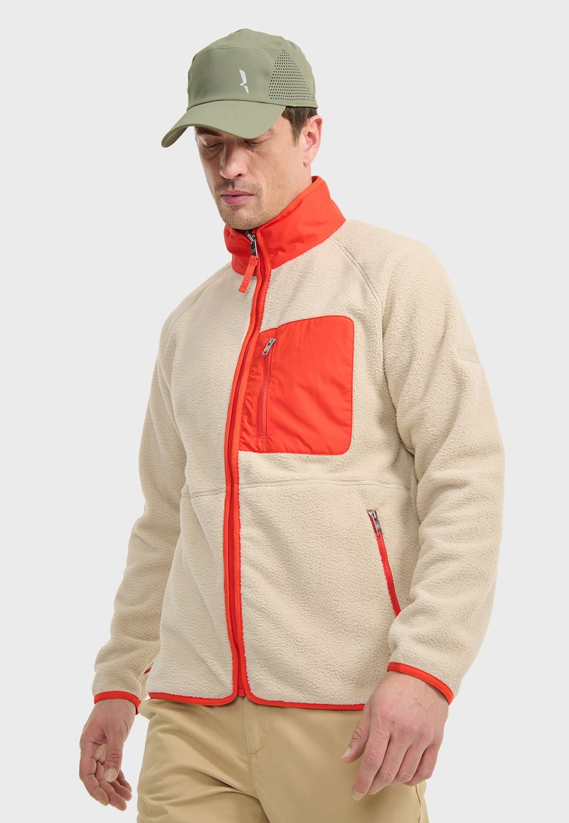 Veste en polaire crème avec un col rouge et une poche sur la poitrine. Comprend des zips, des coutures contrastées et une coupe décontractée. Accentuée par une casquette en vert olive.