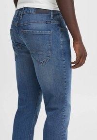 Jeans de denim en azul oscuro con una textura suave, que presentan dos bolsillos traseros, detalles de costura sutiles y una etiqueta de cuero de la marca en la cinturilla.
