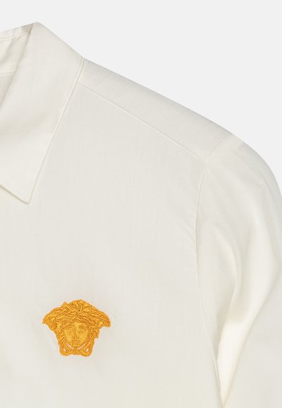 Camicia bianca con colletto, con un logo di Medusa ricamato in oro sul lato sinistro del petto, tessuto liscio con una leggera lucentezza e cuciture pulite.