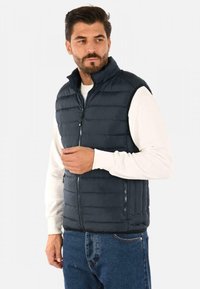 Gilet imbottito navy, con colletto alto, chiusura con zip e tasche laterali. Indossato sopra una camicia bianca a maniche lunghe e abbinato a jeans blu.
