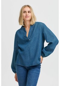 Top in denim con scollatura a V e maniche lunghe arricciate. Il tessuto è di un blu medio, con una vestibilità morbida e dettagli di cucitura minimi.