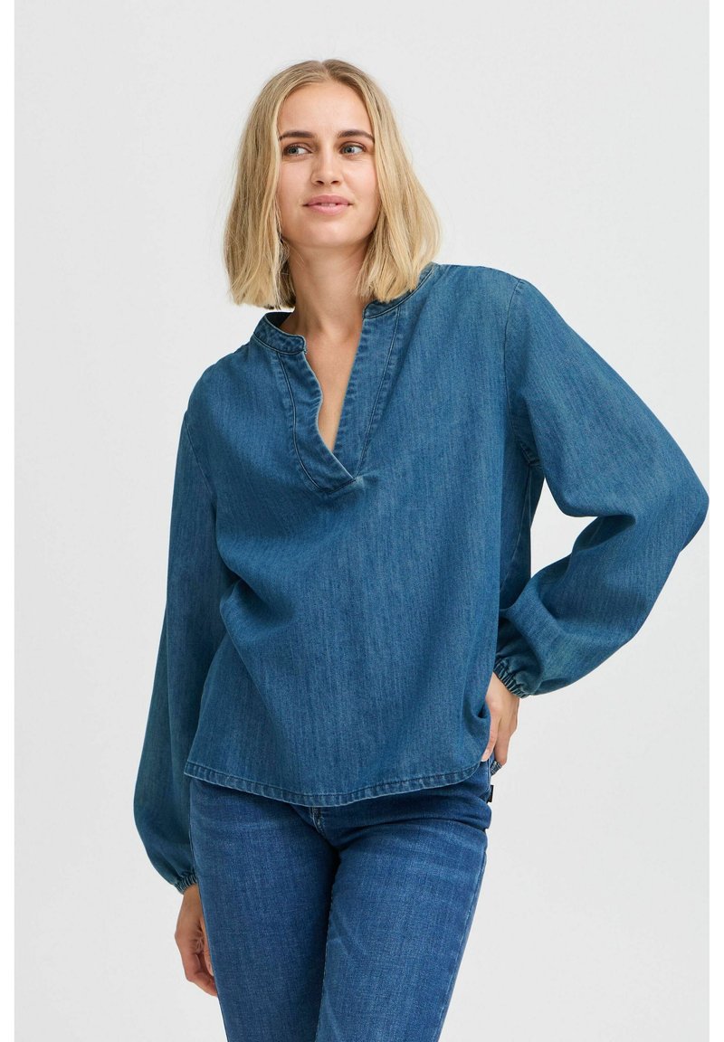 Top in denim con scollatura a V e maniche lunghe arricciate. Il tessuto è di un blu medio, con una vestibilità morbida e dettagli di cucitura minimi.