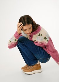 Roze pleteni pulover s cvjetnim uzorkom na prednjem dijelu, u kombinaciji s plavim trapericama i svijetlosmeškim zamščenim cipelama s laganim potplatima.