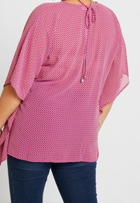 Haut rose à motifs avec manches amples et fluides, doté d'une fermeture par nœud à l'arrière et d'un design texturé en grille. Associé à un jean foncé.