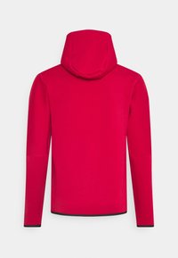 Röd huvtröja med kenguruficka, långa ärmar och dragsko i huvan. Har en kontrasterande svart nederkant och muddar, slät textur.