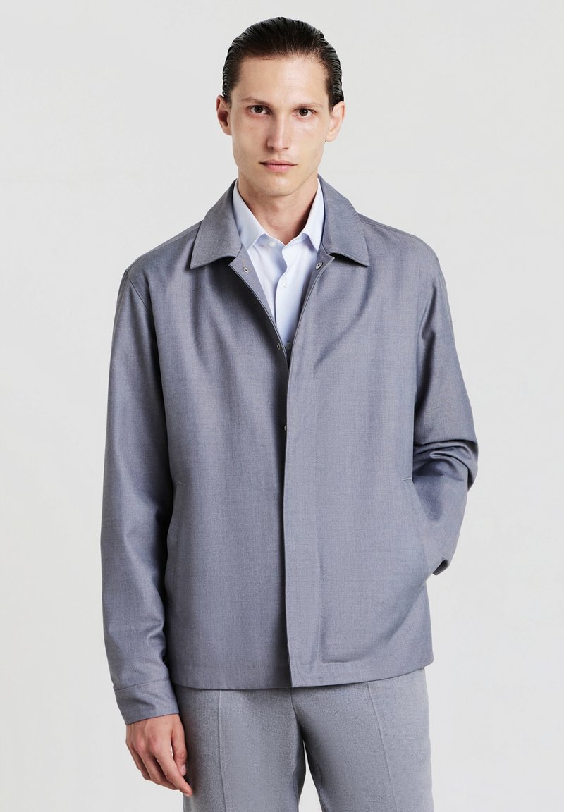 Giacca grigia leggera in un tessuto testurizzato con colletto a punta, chiusura frontale a bottoni a pressione e tasche laterali. Maniche lunghe con un fit rilassato.