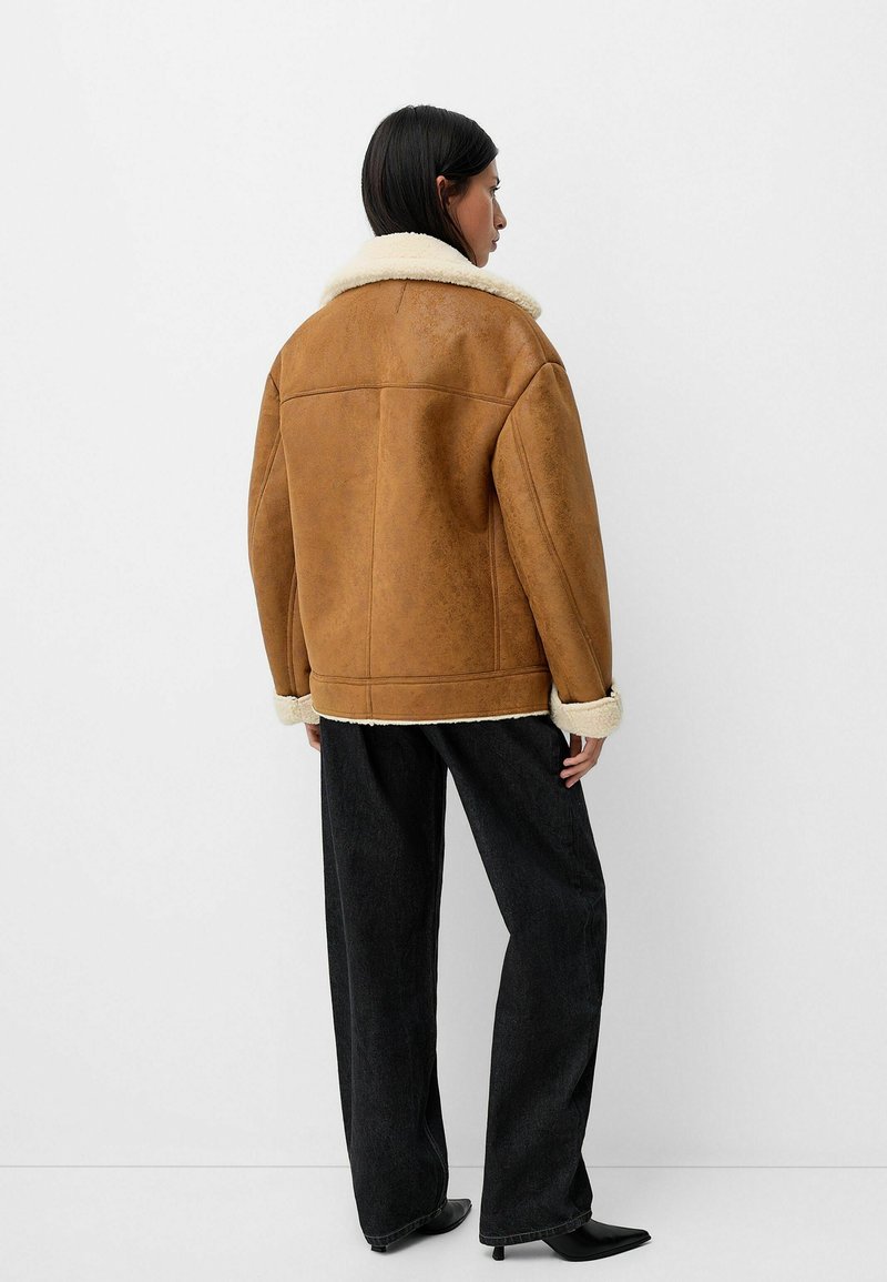 Mujer de espaldas, vestida con una chaqueta de borrego marrón, pantalones anchos negros y tacones de punta negra de estilo kitten, contra un fondo claro y sencillo.