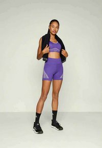 Γυναίκα που στέκεται με μοβ αθλητικό σουτιέν και σορτς, κρατώντας μια μαύρη πετσέτα στους ώμους, φορώντας μαύρα αθλητικά παπούτσια και κάλτσες, μπροστά σε λευκό φόντο.