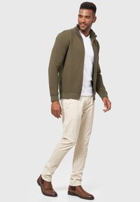 Chaqueta de punto verde oliva con cuello alto, combinada con una camiseta blanca, pantalones beige y botas de tobillo marrones. Detalle de tela texturizada.