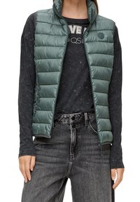 Vadderad grön väst med hög krage och dragkedjestängning. Texturerad tyg med horisontella sömmar. Bärs över en svart långärmad skjorta och grå jeans.