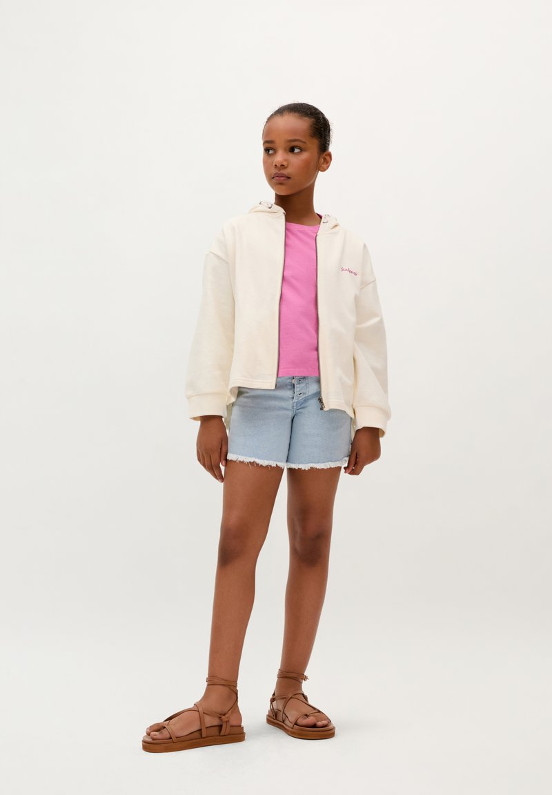 Lichtgekleurde crème zip-up hoodie, kort roze t-shirt, denim shorts met rafelige zoom, en bruine sandalen met bandjes. Eenvoudige, casual outfit.